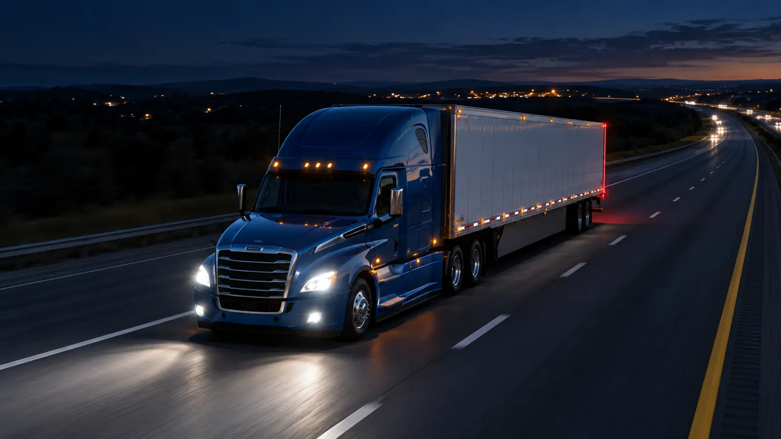 Blue Class A tractor-trailer operating with headlights and marker lights on a divided interstate at night